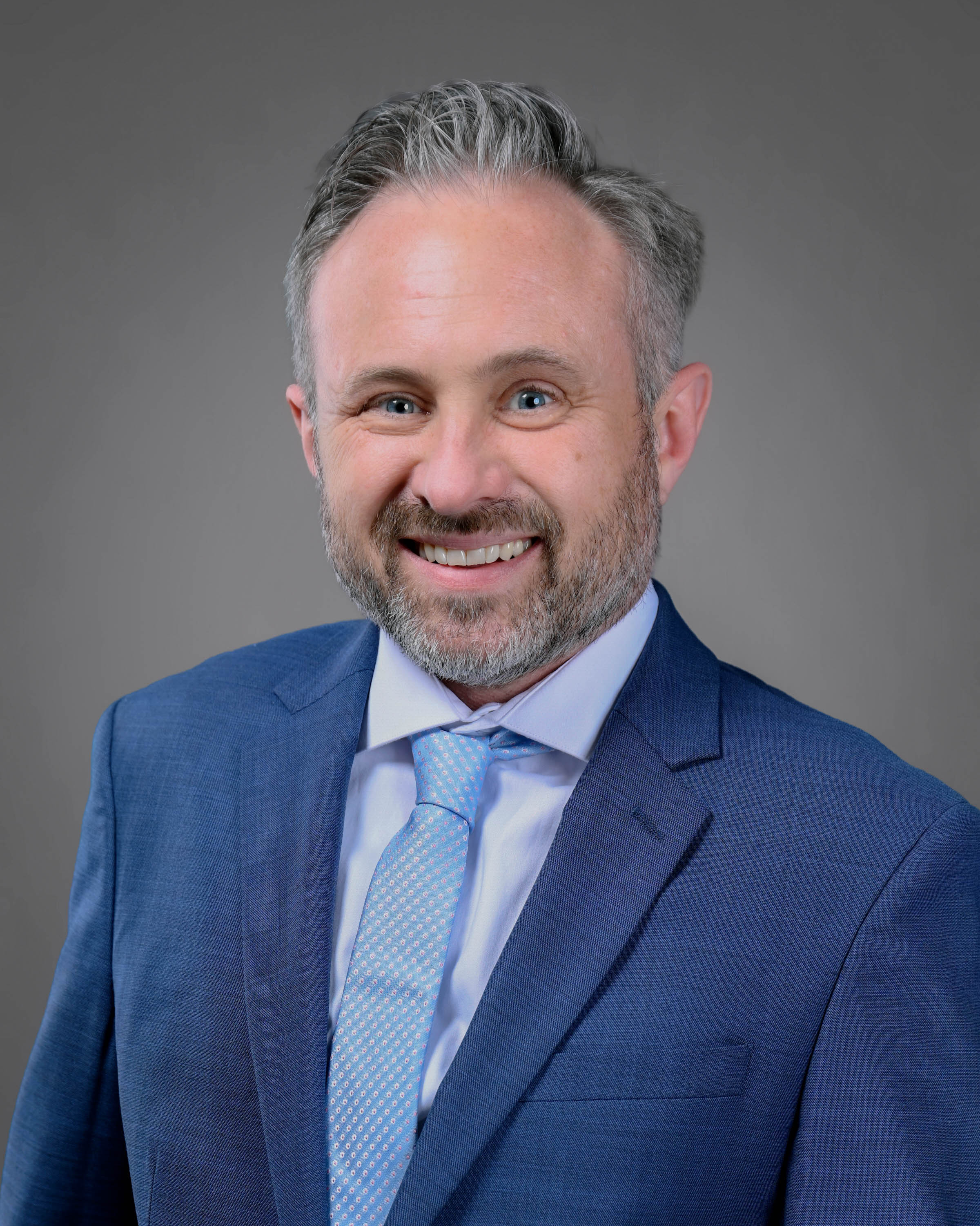 Smiling man with dark and silver hair and a short beard in a blue suit with a lighter blue shirt and tie.